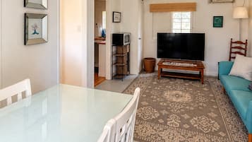 A living room with a glass-top dining table, white chairs, a sofa, a television, a ceiling fan, and a patterned rug.