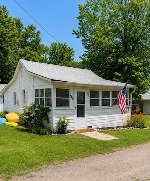 A single-story house with a gabled roof, a front porch, and a small front yard with a flag. - Plum Point Cottage #17 (Huntingtown)