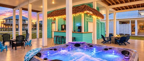 A hot tub with a wooden deck, outdoor seating, and a tiki bar.