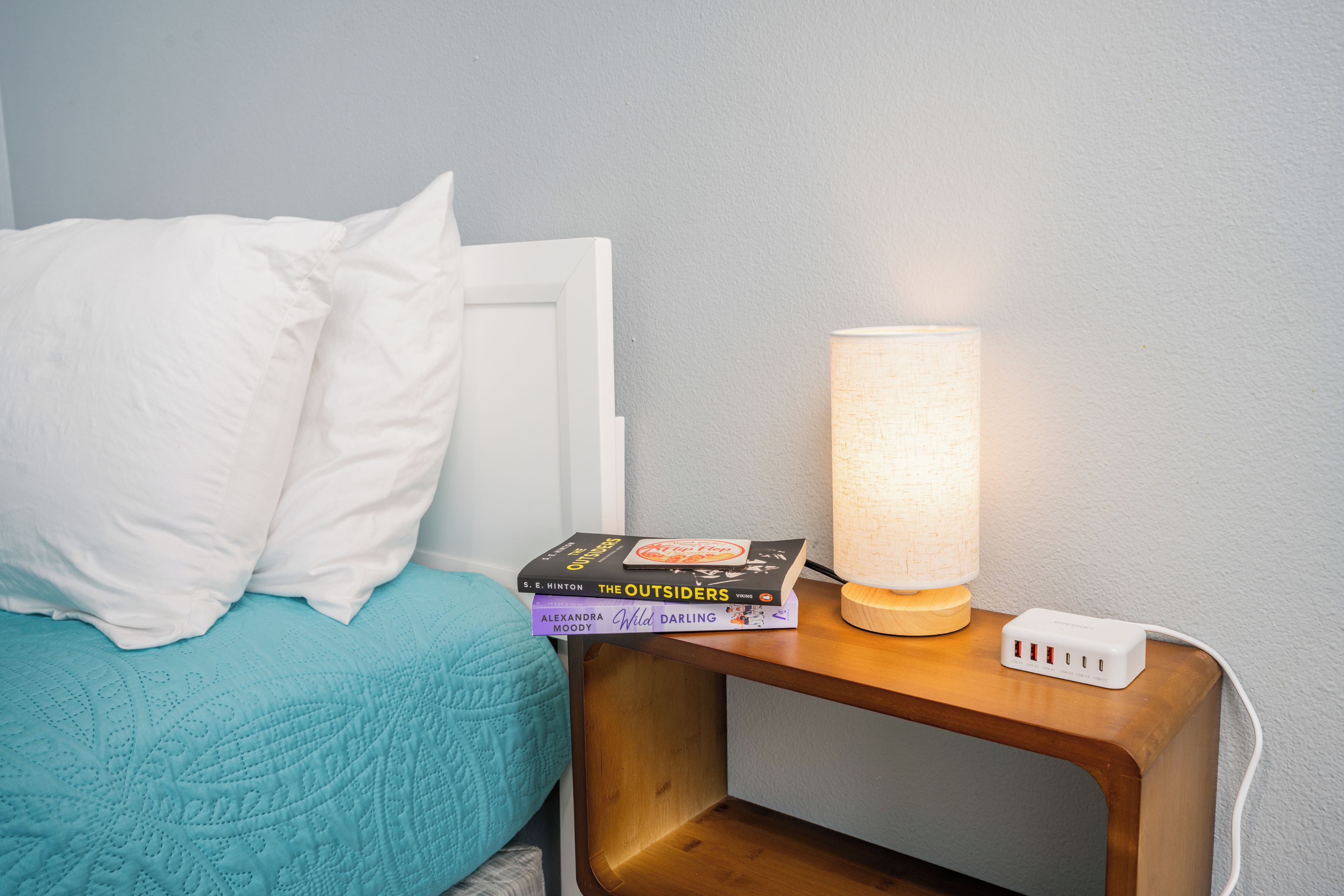 Una cama con almohada blanca y manta verde azulada, una mesita de noche con lámpara, y una pila de libros.