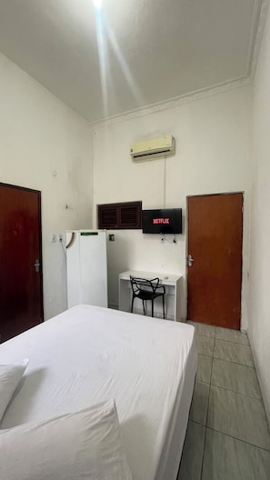 Quarto de hotel com uma cama, uma escrivaninha com cadeira, uma televisão e um ar condicionado.