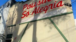 Fachada de edifício com uma placa grande escrita 'Pousada So Alegria' em letras vermelhas.