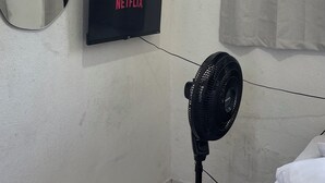Um quarto com uma cama, uma TV fixada na parede exibindo 'NETFLIX', um ventilador e uma janela com vista para edifícios.