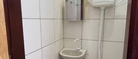 Une petite salle de bain carrelée, équipée de toilettes, d’un lavabo et d’un porte-rouleau de papier toilette fixé au mur.