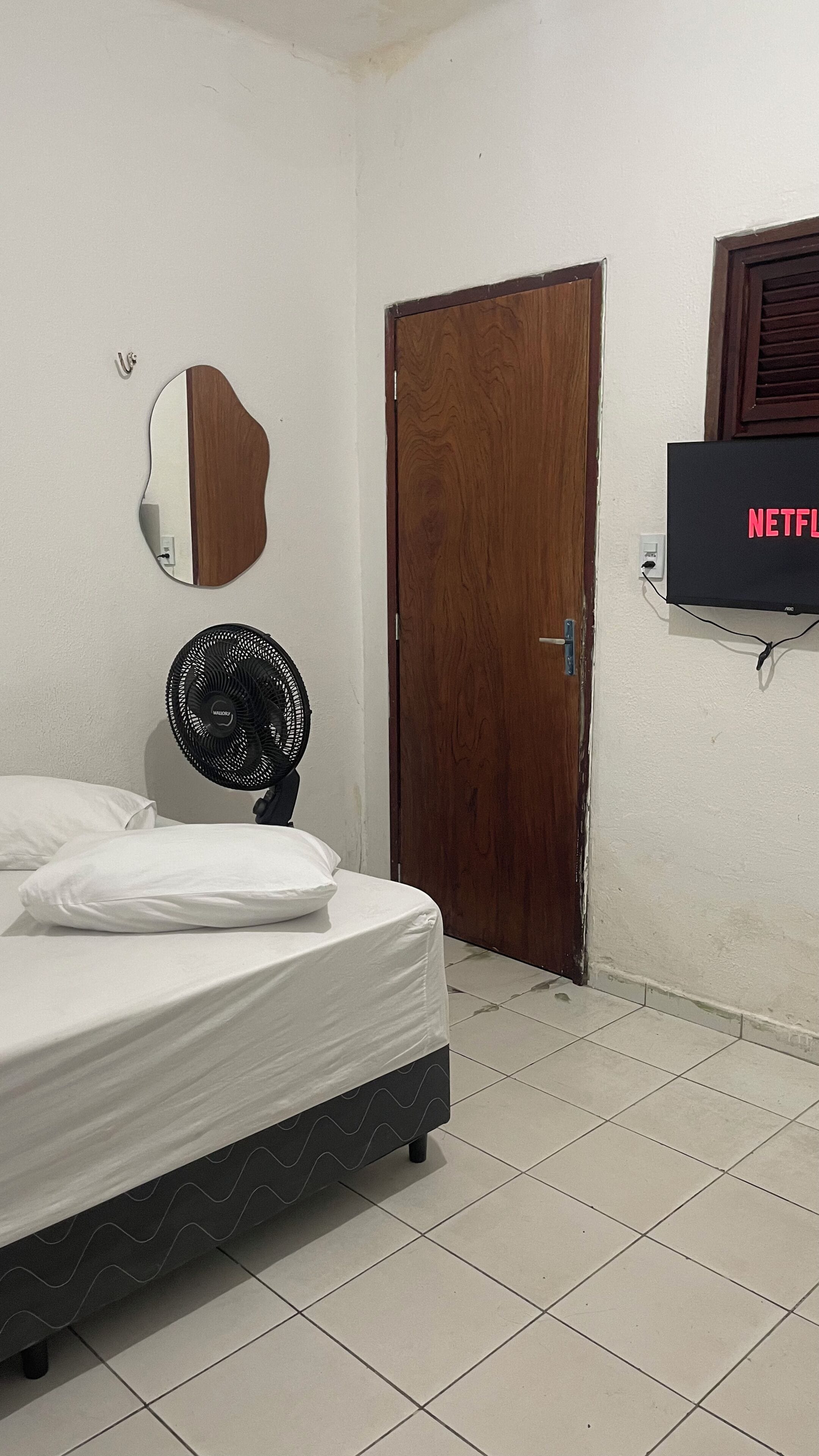 A bedroom with a bed, a wooden door, a wall-mounted TV, and a standing fan.