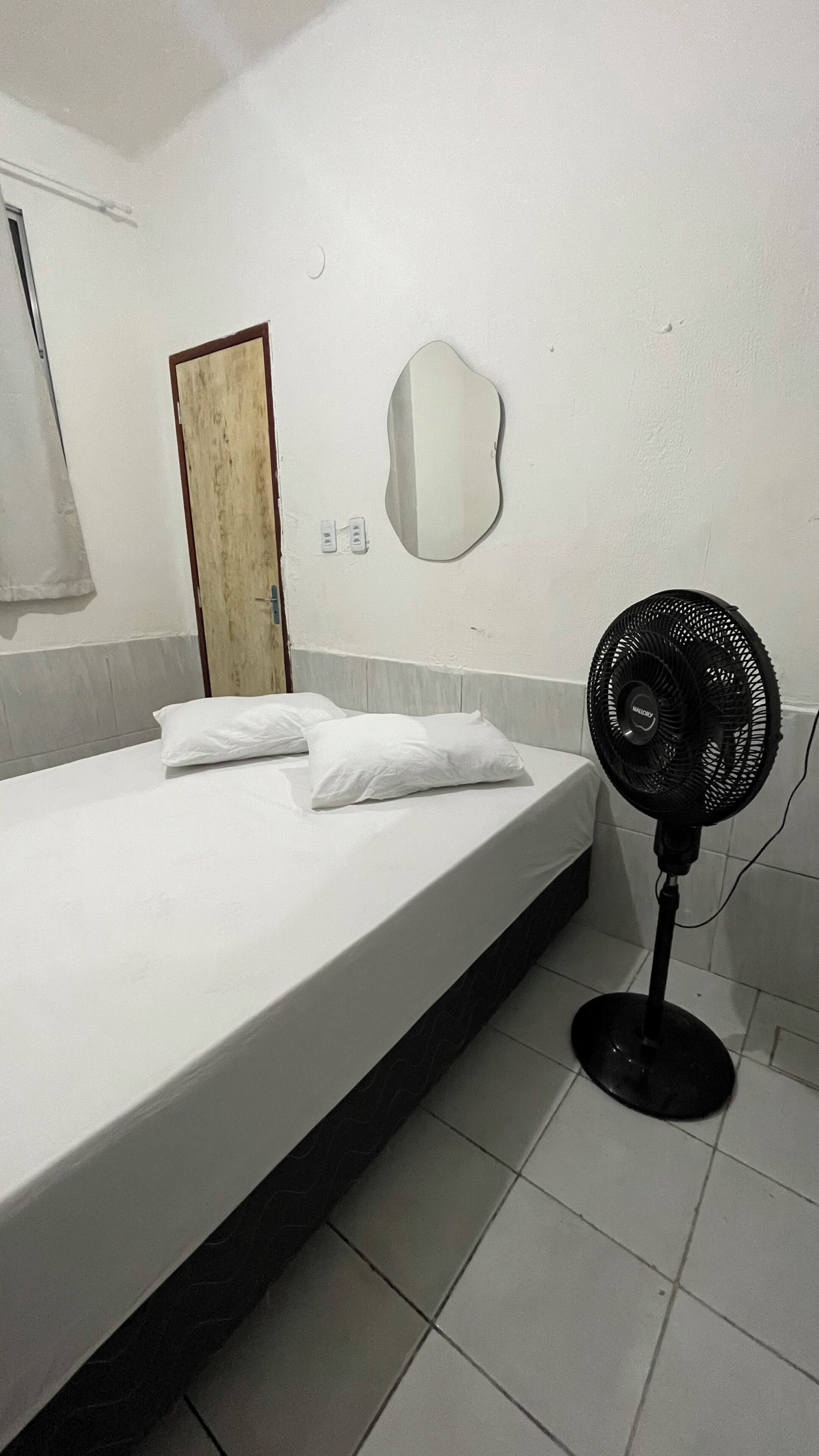 Habitación de hotel con cama, almohadas, espejo fijado a la pared, ventilador de pie y una puerta.