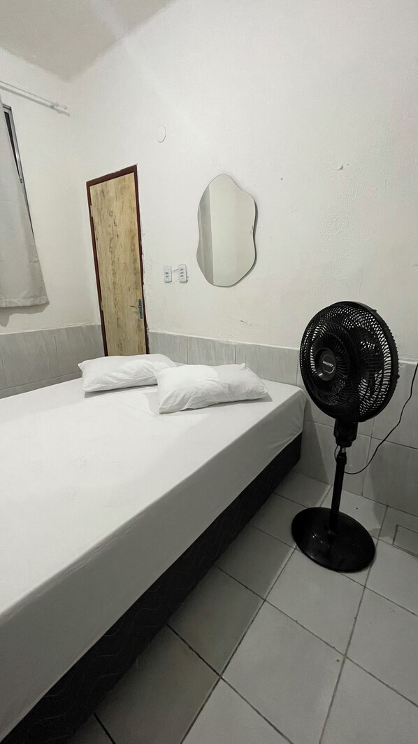 Habitación de hotel con cama, almohadas, espejo fijado a la pared, ventilador de pie y una puerta.