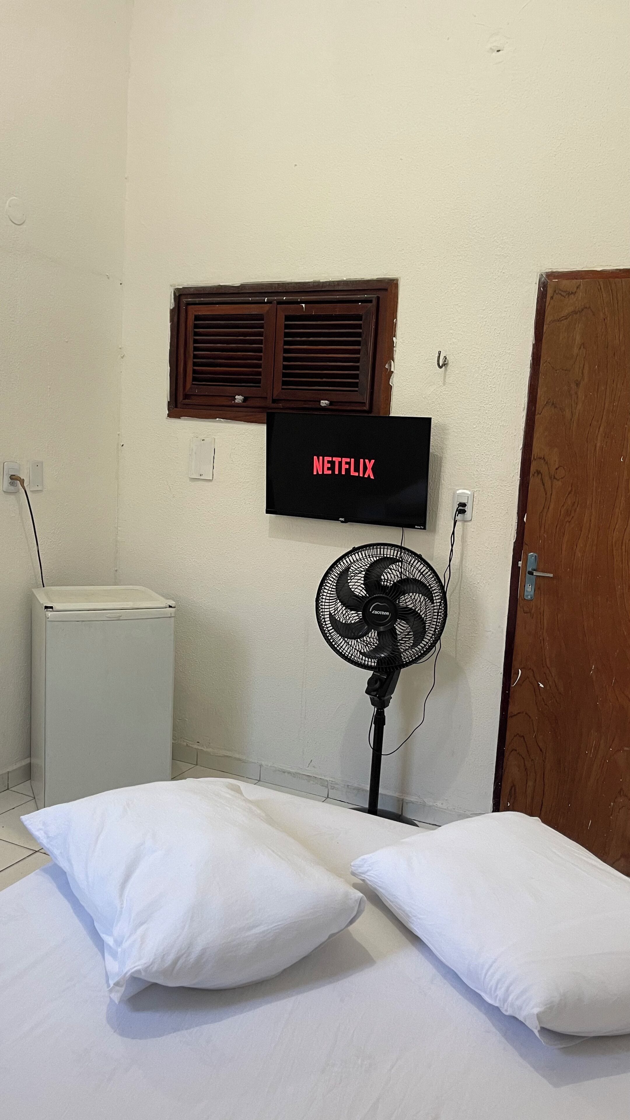 Ein Zimmer mit einem Bett, einem an der Wand befestigten Fernseher, der 'NETFLIX' anzeigt, einem Standventilator und einem Fenster mit Holzjalousien.