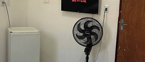 Ein Zimmer mit einem Bett, einem an der Wand befestigten Fernseher, der 'NETFLIX' anzeigt, einem Standventilator und einem Fenster mit Holzjalousien.