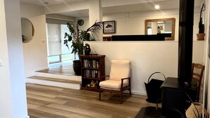 Uma sala de estar com piso de madeira, uma estante de livros, uma cadeira, um espelho e um vaso com planta.