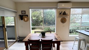 A dining area with a wooden table, chairs, a potted plant, and a view through large windows.