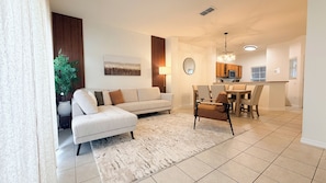 A modern living room with a sofa, a dining table with chairs, a kitchen area, and a large potted plant. - 3-bedroom Disney Dream Vacation Home with private pool (Kissimmee)