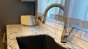 Une cuisine dotée d’un plan de travail en granit, d’un évier en acier inoxydable et d’une fenêtre avec des stores.