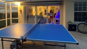 Een tafeltennistafel in een buitenruimte met een glazen omheining, een fiets en een huis op de achtergrond.