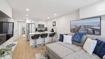 A modern apartment with a kitchenette, a dining area, a living room with a sofa, and a large window with a view.