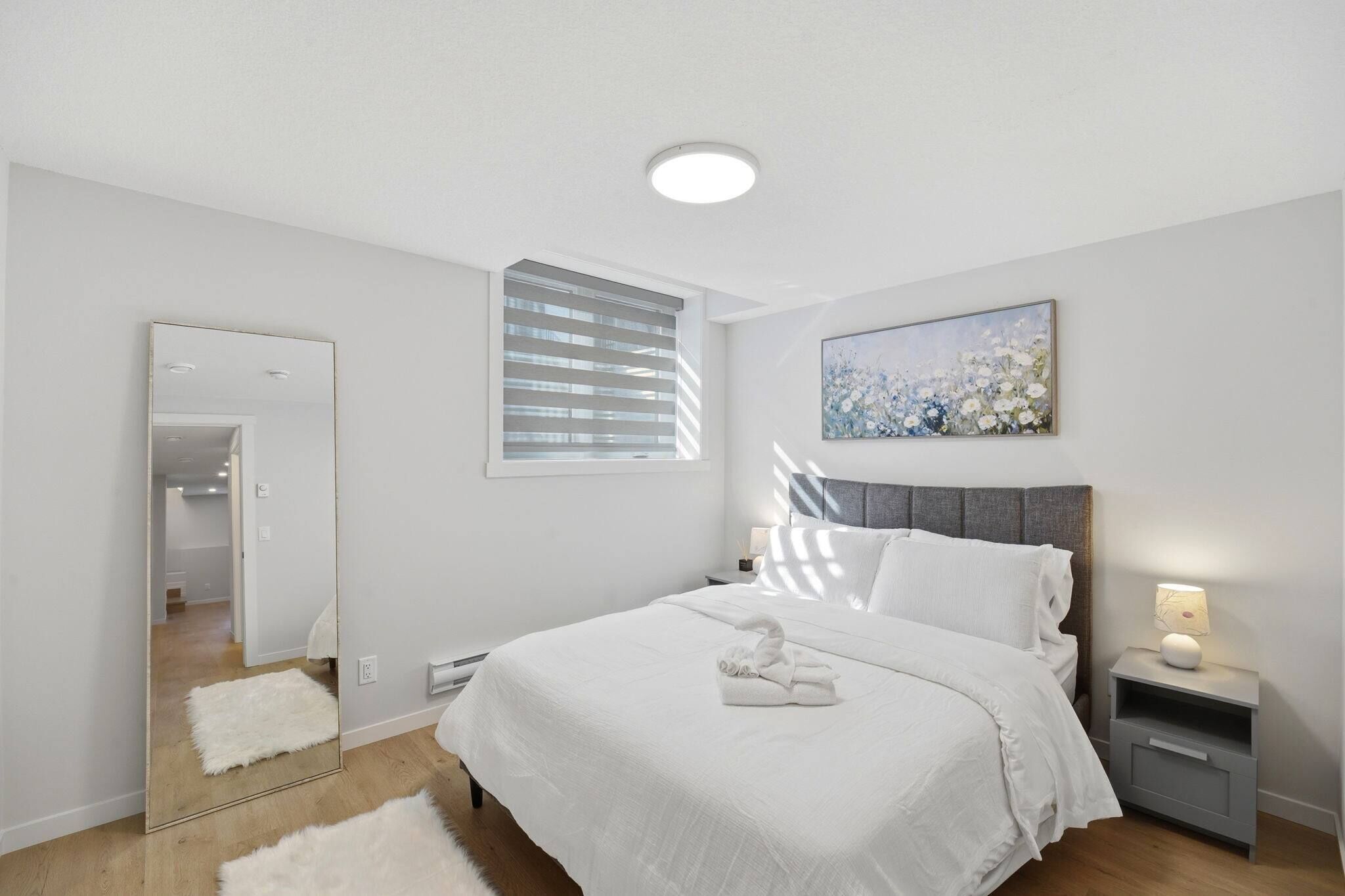 A modern bedroom with a large bed, a bedside table with a lamp, a mirror, and a window with blinds.