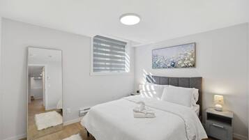 A modern bedroom with a large bed, a bedside table with a lamp, a mirror, and a window with blinds.