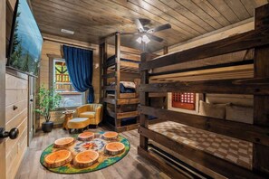 Ein gemütliches Blockhäuschen-Interieur mit Etagenbett, Deckenventilator, Fenster mit Vorhängen, einem kleinen Tisch und einer Pflanze.
