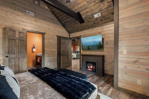 Ein Interieur einer Blockhütte mit Kamin, einem Bett mit dunkler Decke und einem Bild an der Wand.