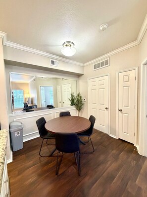 A dining area with a round table and chairs, a trash bin, and a view into another room.
