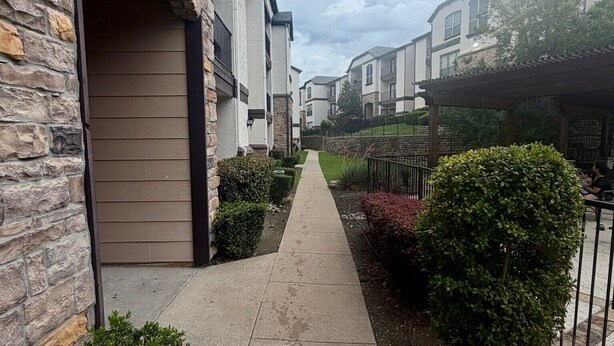 Une rue résidentielle bordée de bâtiments en pierre et en bois, avec un trottoir pavé et des haies bien entretenues.