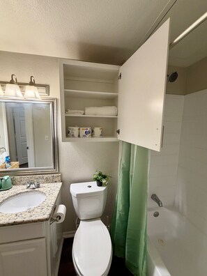 A bathroom with a toilet, sink, mirror, shelves with toiletries, and a shower curtain.