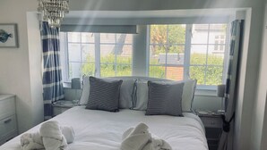 Una habitación de hotel con una cama grande, dos mesitas de noche, un tocador, una araña y una ventana con cortinas.