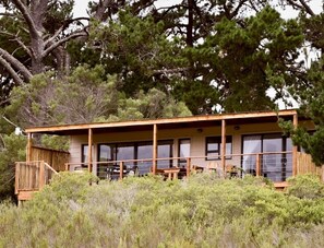 A cabin with a covered porch, large windows, and a view of surrounding trees. - Equleni Farm (Knysna)