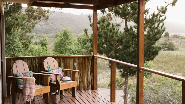 A wooden deck with two chairs and a table, a hat, and a glass on the table. - Equleni Farm (Knysna)