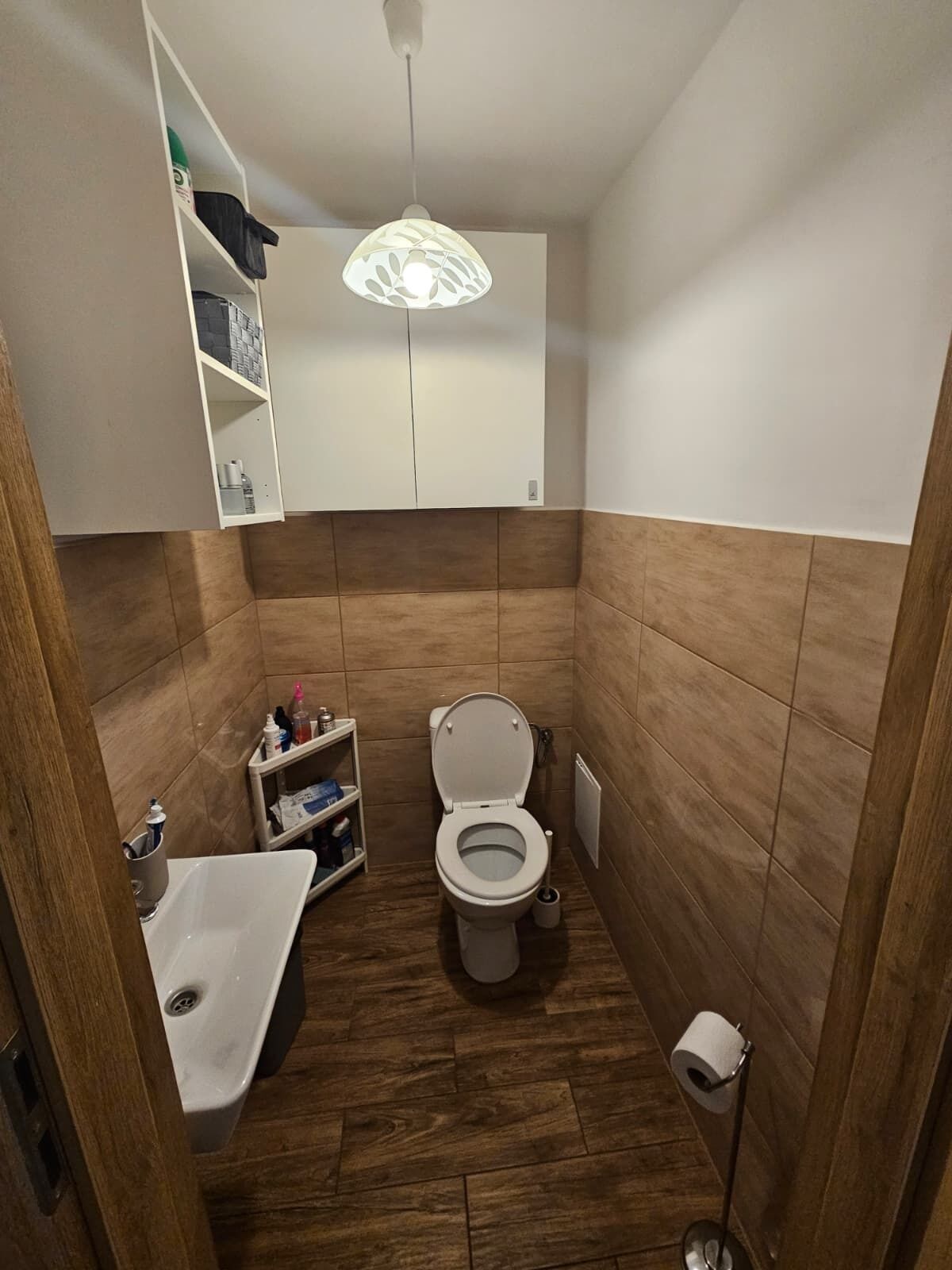 Une petite salle de bain comprenant des toilettes, un lavabo, une étagère avec des articles de toilette et un luminaire suspendu.