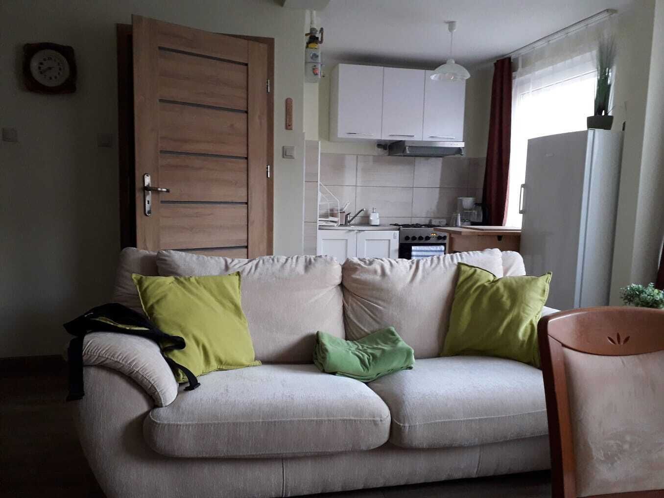 A living room with a sofa, a wooden door, a kitchen area with cabinets and appliances, and a window with curtains.