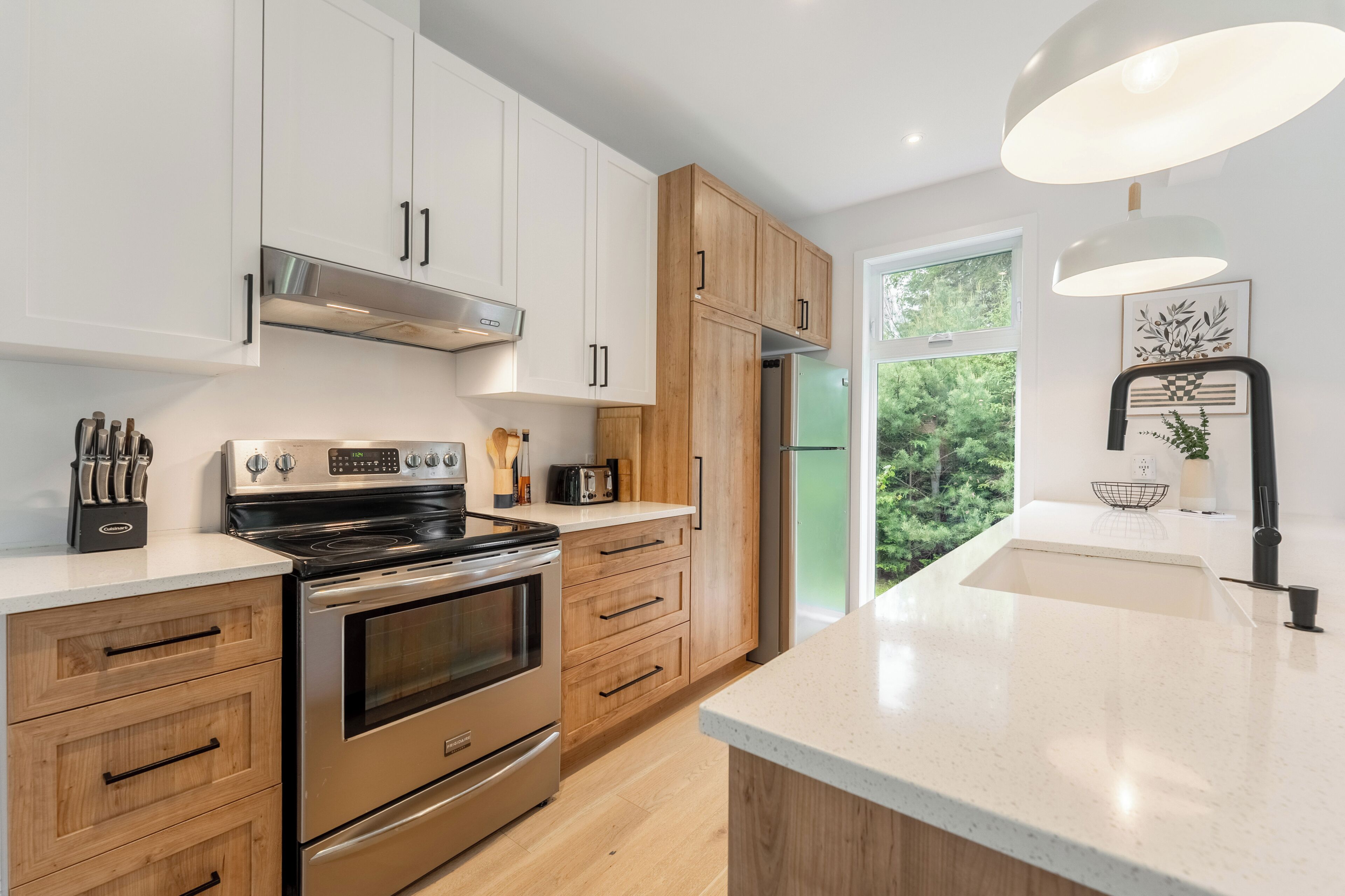 A modern kitchen with stainless steel appliances, wooden cabinets, and a large window providing a view of greenery.