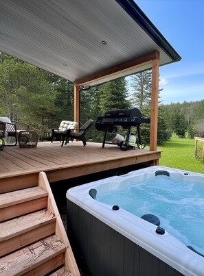 Een houten terras met een jacuzzi, tuinmeubilair en een barbecue.