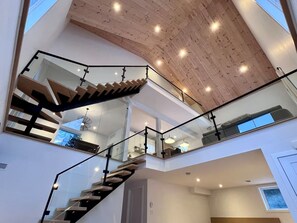 Ein modernes Interieur mit einer Treppe, die zu einem Erker führt, Glasgeländern und einer Holzbalkendecke.