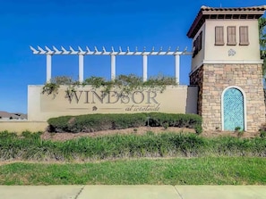 Schild für 'Windsor at Westside' mit einer Pergola darüber, einem Steinhaus mit blau gefliestem Fenster und gepflegter Bepflanzung.