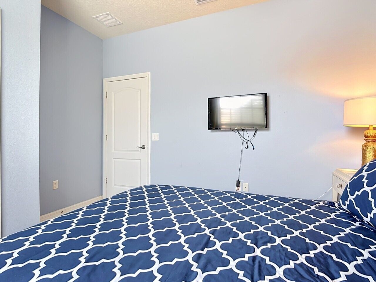 Ein Schlafzimmer mit einem Bett, einem an der Wand befestigten Fernseher, einer Tür und einem Nachttisch mit Lampe.