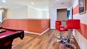 Uma mesa de bilhar em um espaço com piso de madeira e cadeiras vermelhas.