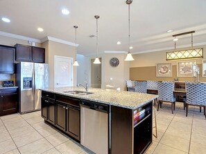 A modern kitchen with a central island, stainless steel appliances, pendant lighting, and a dining area with patterned chairs. - Windsor Westside 8BR Lake View Home Near Disney (Kissimmee)