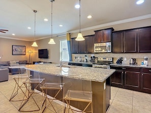 A modern kitchen with dark wood cabinets, a granite countertop, a built-in microwave, and a large island with bar stools. - Windsor Westside 8BR Lake View Home Near Disney (Kissimmee)