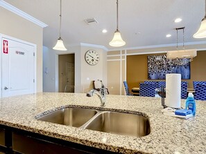 A kitchen with granite countertops, stainless steel sinks, pendant lighting, a clock, and a dining area with blue upholstered chairs. - 8BR Cozy Pool Villa, Lake View, Water Slides (Kissimmee)
