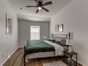 Un dormitorio con cama, ventilador de techo, ventana con persianas, mesita de noche con lámpara y estante junto a la cama con libros.