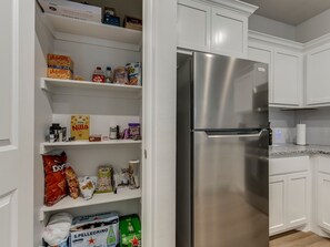 Una cocina con armarios blancos, un refrigerador de acero inoxidable y estantes llenos de alimentos.