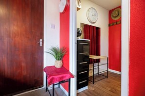 Um corredor com porta de madeira, parede vermelha, uma mesa pequena com planta, armário preto e um relógio na parede.