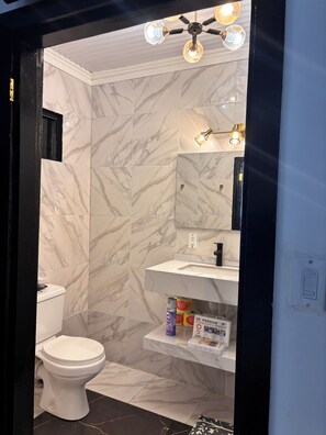 Une salle de bain avec des toilettes blanches, des carreaux de marbre, un ventilateur de plafond et une étagère contenant des articles de toilette.