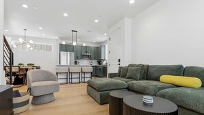 Een moderne woonkamer met een grijze bank, een poef en een zwarte salontafel. Op de achtergrond is een trap met een leuning te zien.