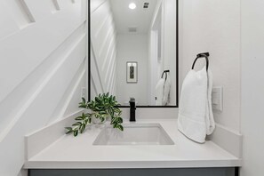 A modern bathroom with a white sink, a black faucet, a potted plant, a white towel, and a framed picture on the wall.