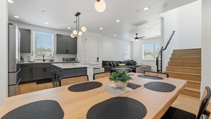 Een moderne keuken met grijze kastjes, een eethoek met een houten tafel en zwarte matten, een trap en een woongedeelte met een bank en een plant.