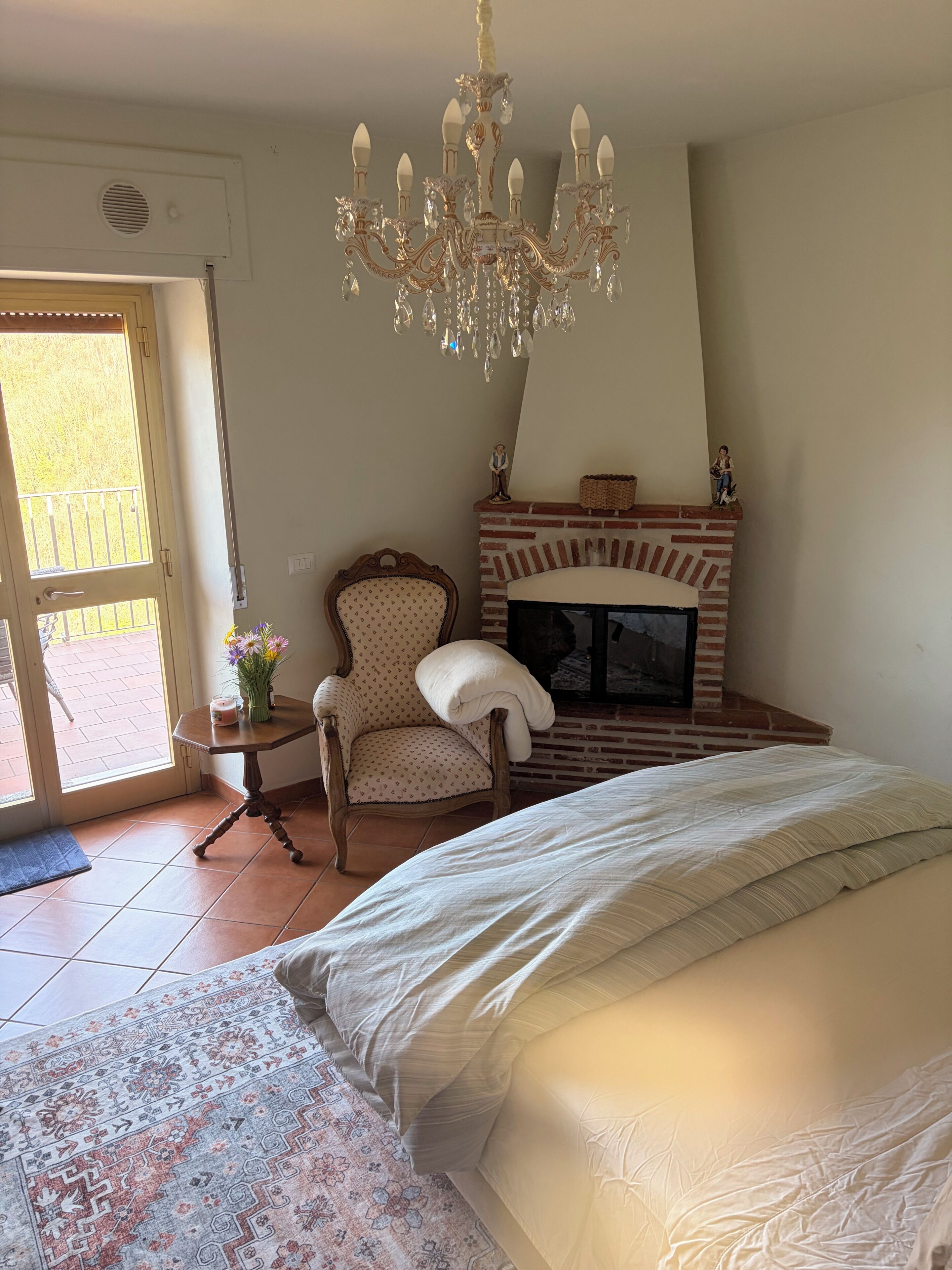 A bedroom with a large bed, a chandelier, a fireplace, a chair, and a small table with flowers.