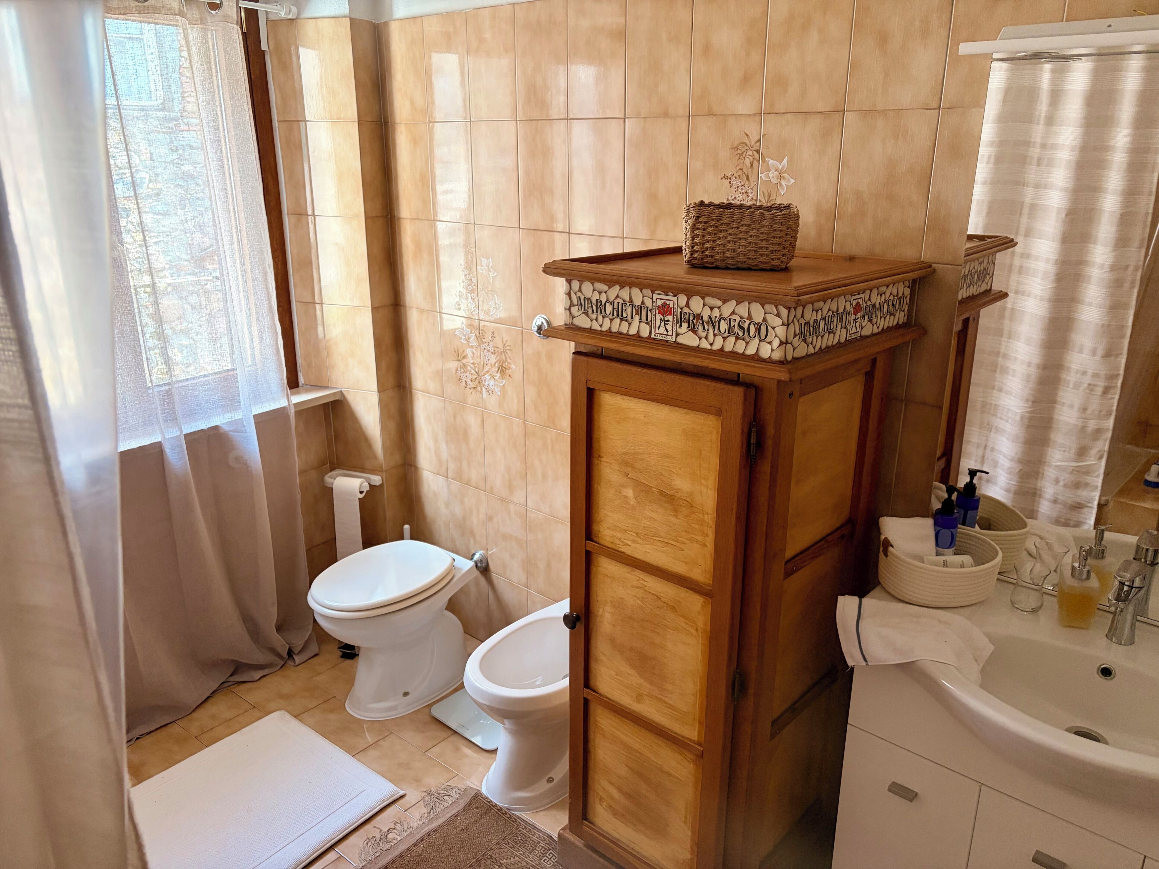 Un baño con dos inodoros, un armario de madera con una cesta, un lavabo con artículos de aseo y una pared con azulejos.