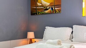 Ein Hotelzimmer mit einem Bett, weißen Laken, einem Sonnenuntergangsbild mit Palmen und einer Lampe.